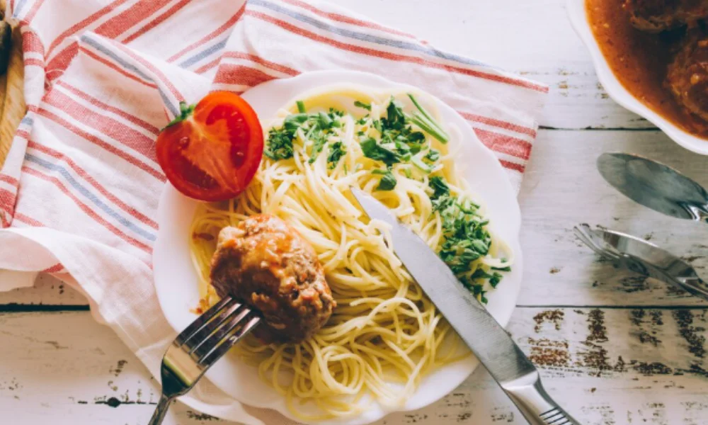 A plate of spaghetti with a meatball, topped with sauce and herbs, sits on a rustic wooden table