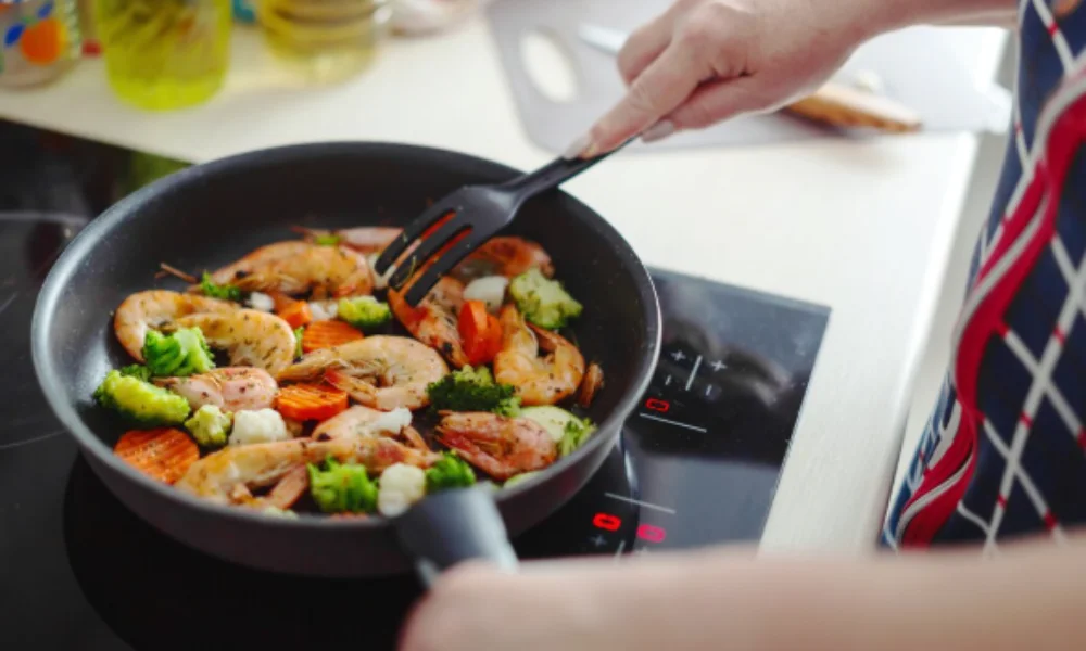 A person cooks shrimp, carrots, and broccoli in a black pan on an induction stove, using a spatula-easy shrimp scampi recipe