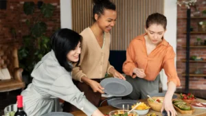Three women happily set a dining table with plates and a variety of colorful dishes,