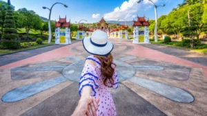 A woman in a pink dress and white hat is leading the viewer down a pathway toward a traditional temple
