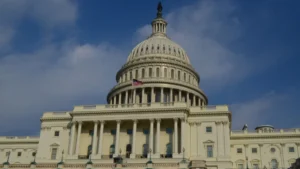 The image shows the United States Capitol building under a blue sky with clouds