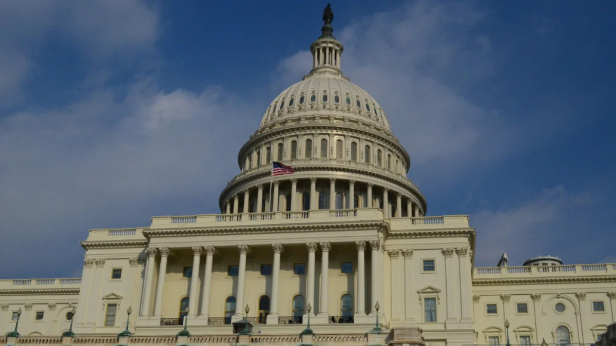 The image shows the United States Capitol building under a blue sky with clouds