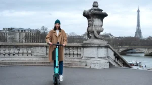A woman in a tan coat and teal beanie rides a teal e-scooter on a Parisian bridge
