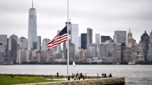 American flag at half-mast against a cloudy sky, with New York City