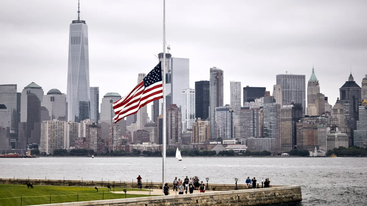American flag at half-mast against a cloudy sky, with New York City
