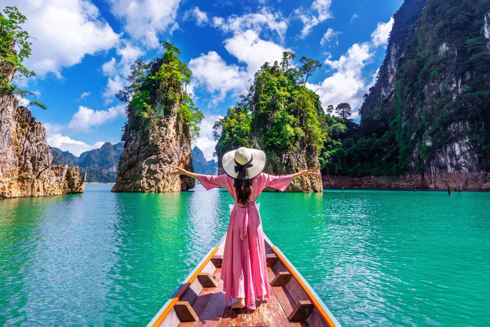 A woman in a pink dress and wide-brimmed hat stands with outstretched arms on a boat