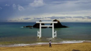 A person stands on a sandy beach facing a large white torii gate partially submerged in the calm ocean