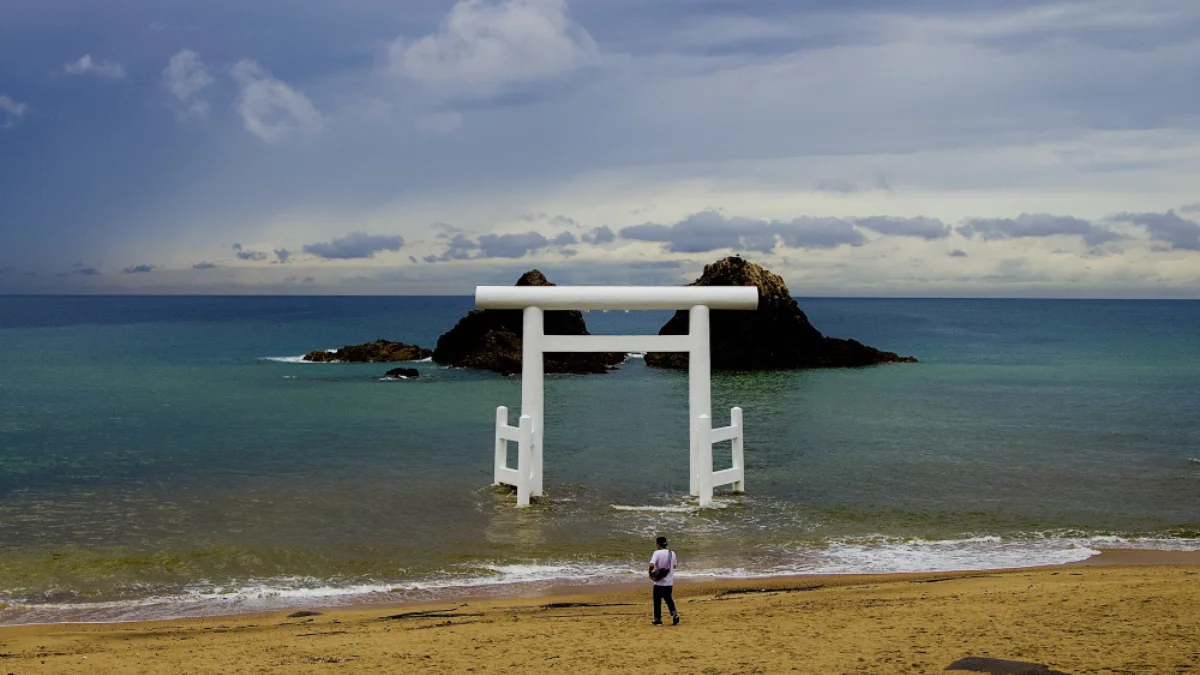 A person stands on a sandy beach facing a large white torii gate partially submerged in the calm ocean