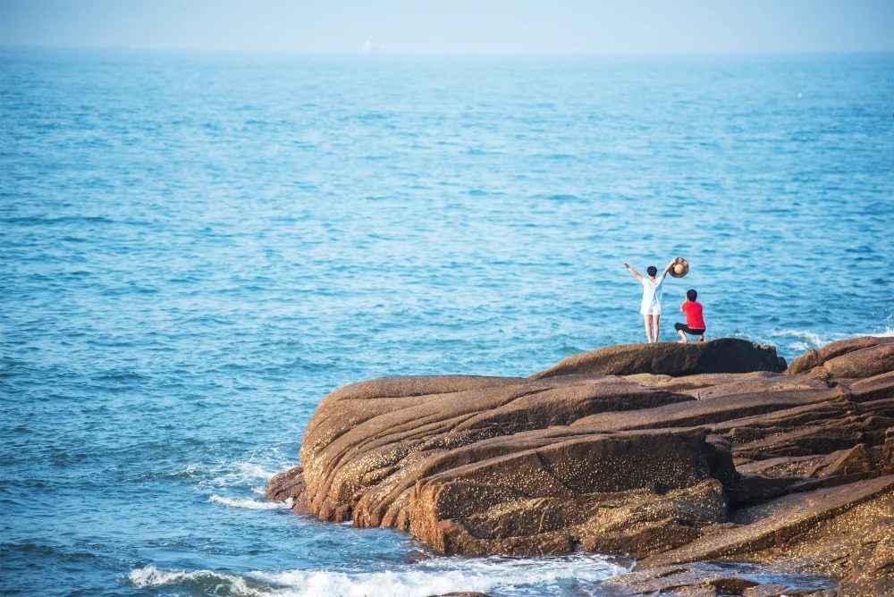 Two people stand on large, sunlit rocks by the sea