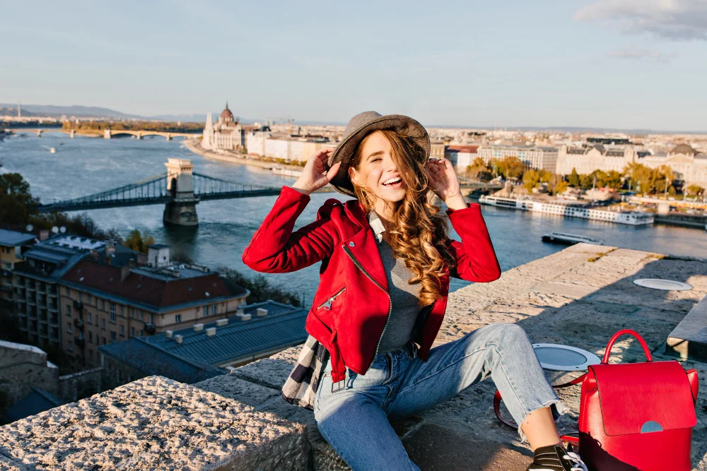 A woman in a red jacket and hat sits on a stone ledge, smiling widely