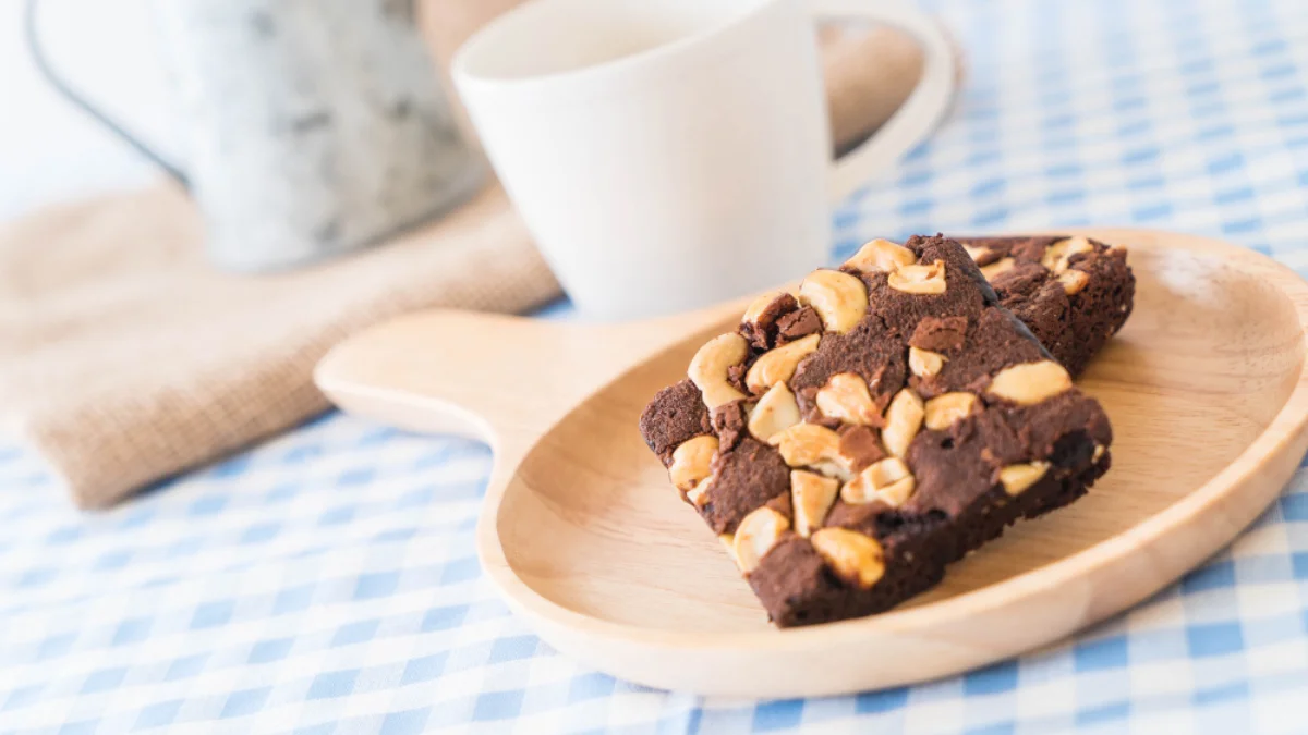 Two brownies topped with cashew nuts sit on a wooden plate with a blue checkered tablecloth
