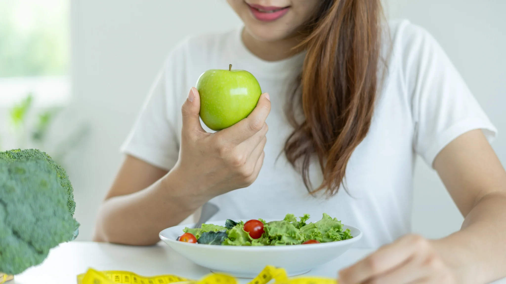 A woman smiles while holding a fresh apple in one hand and a bowl of green salad in the other.