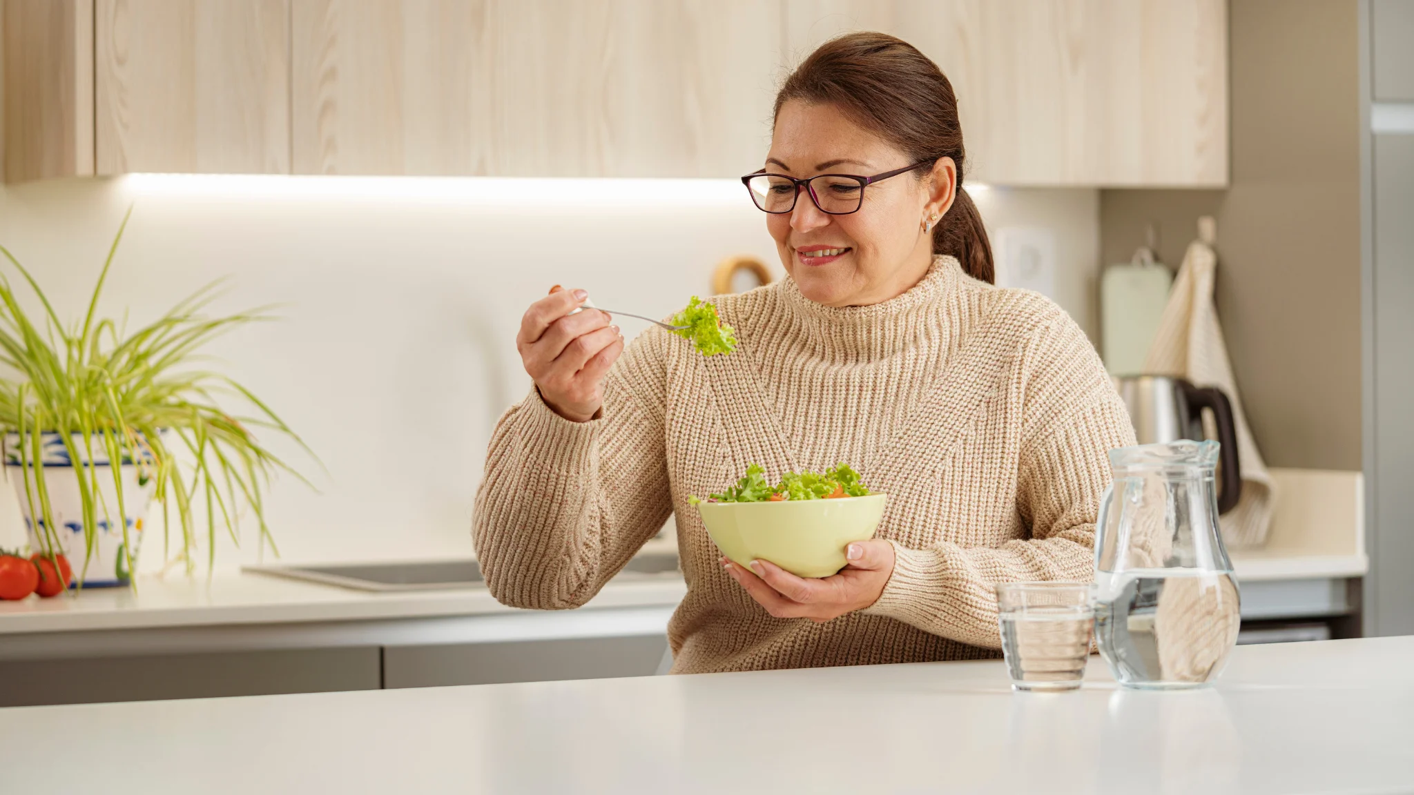 A woman enjoys a fresh salad while sitting at her kitchen table, surrounded by bright, natural light.
