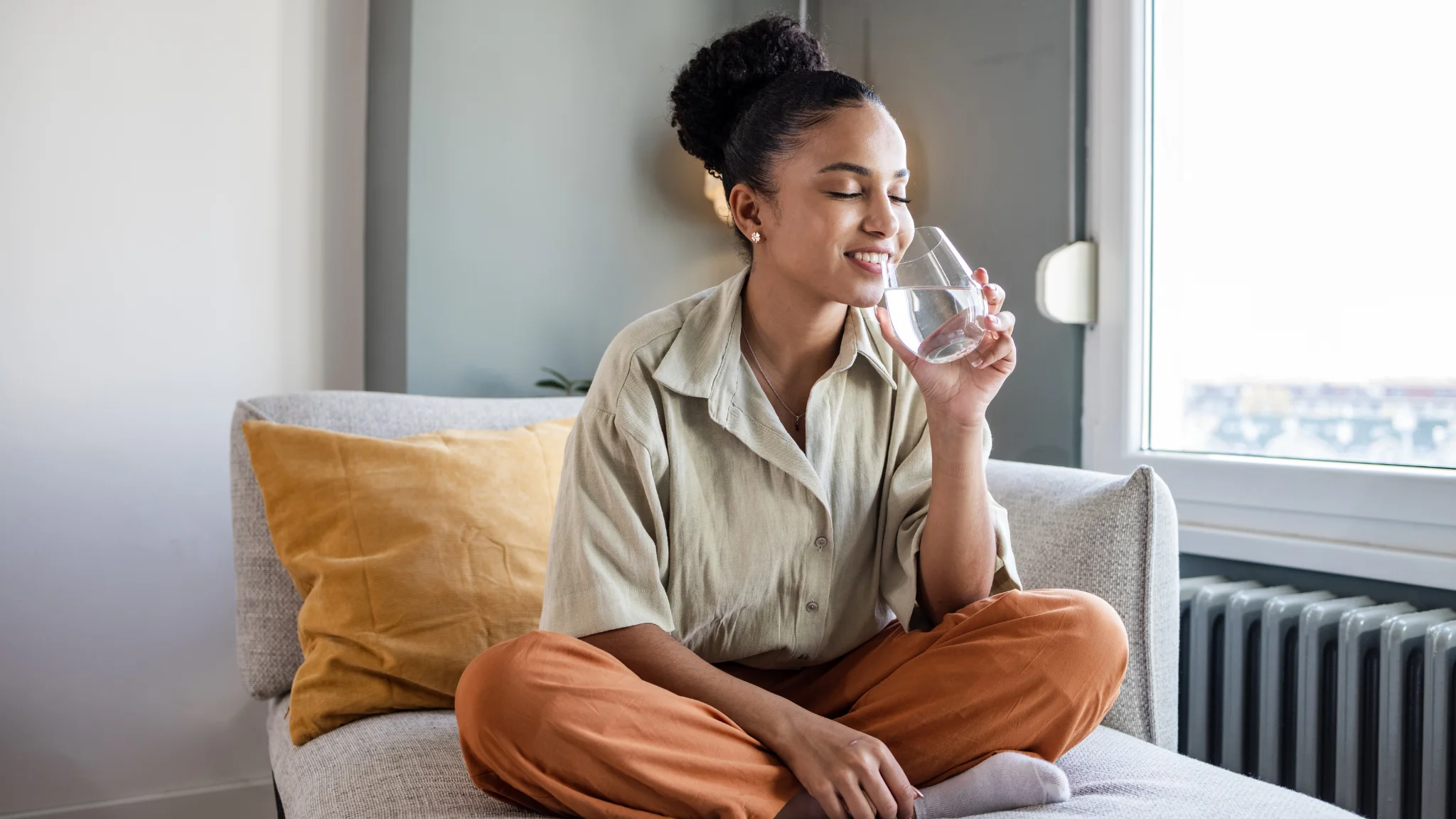 A woman sitting on a couch, sipping water from a glass, looking relaxed and content.