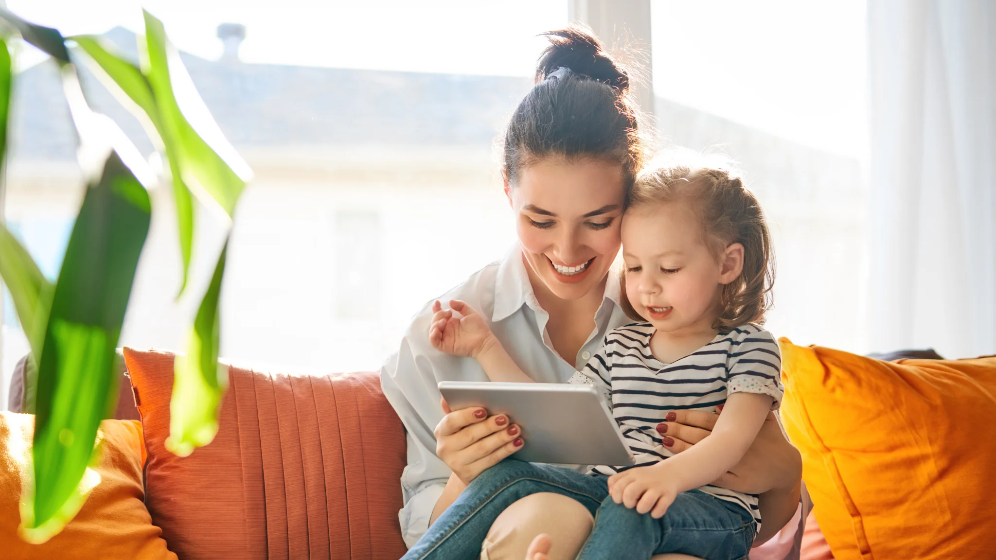 A woman and a child sit together on a couch, both focused on a tablet in their hands.