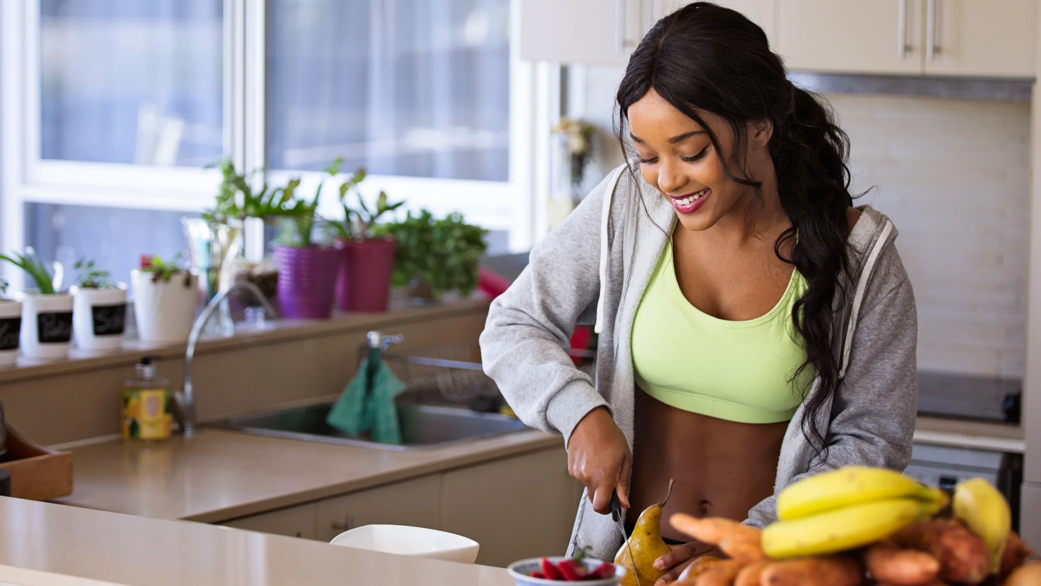 A woman in a green top slices fruit on a kitchen counter, surrounded by fresh produce.