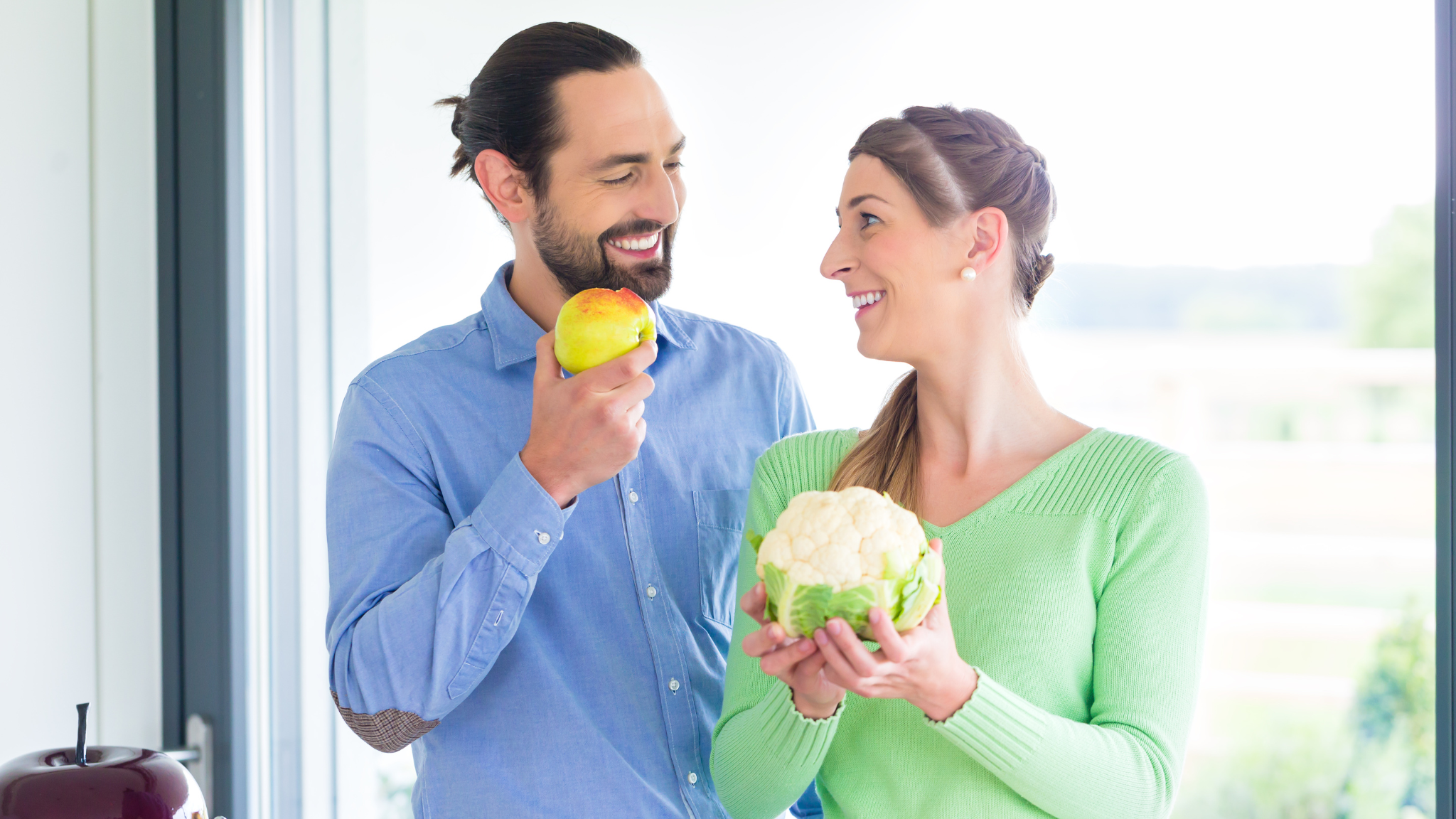 A man and woman smile while holding fresh apples and a bright orange carrot in their hands.