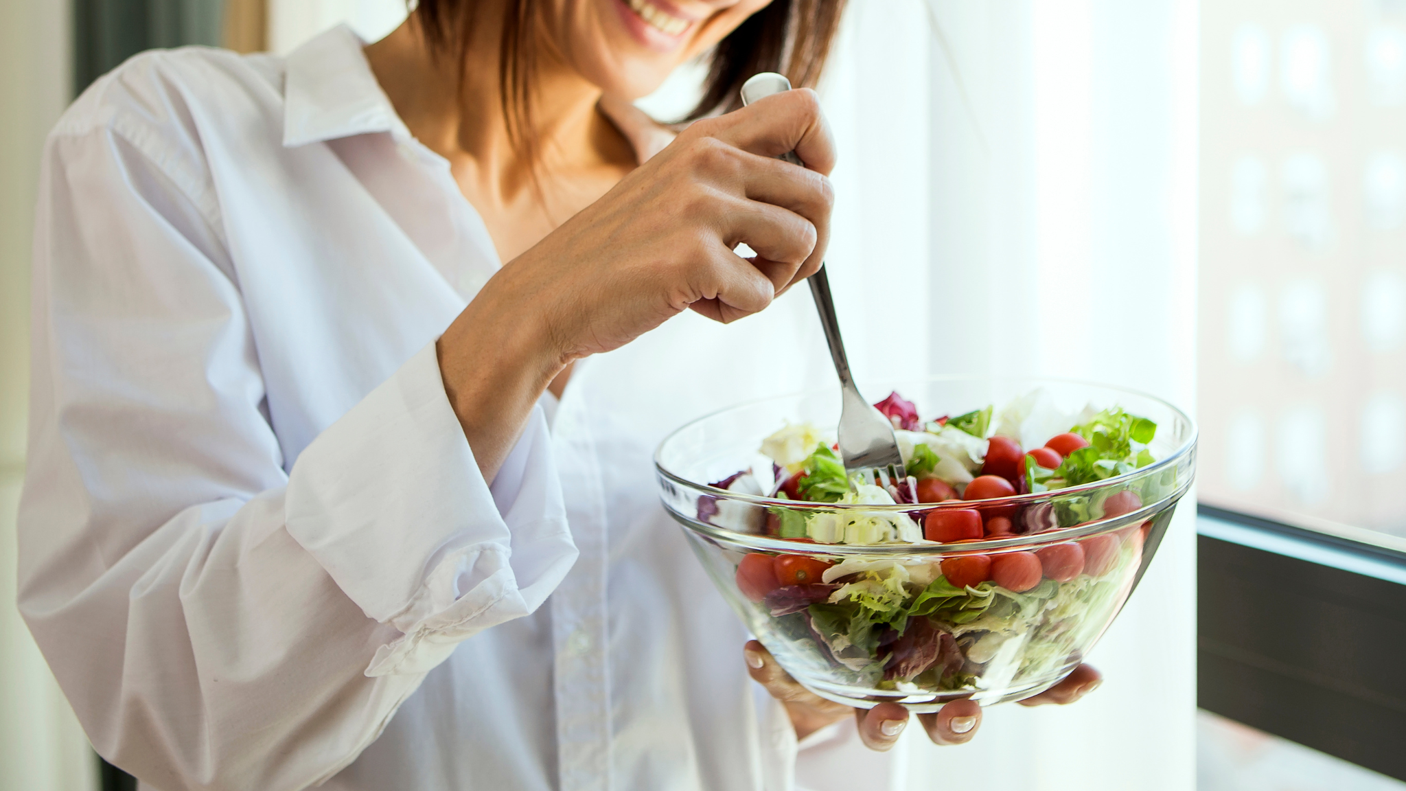 A woman smiles while holding a bowl of salad, standing in front of a bright window.