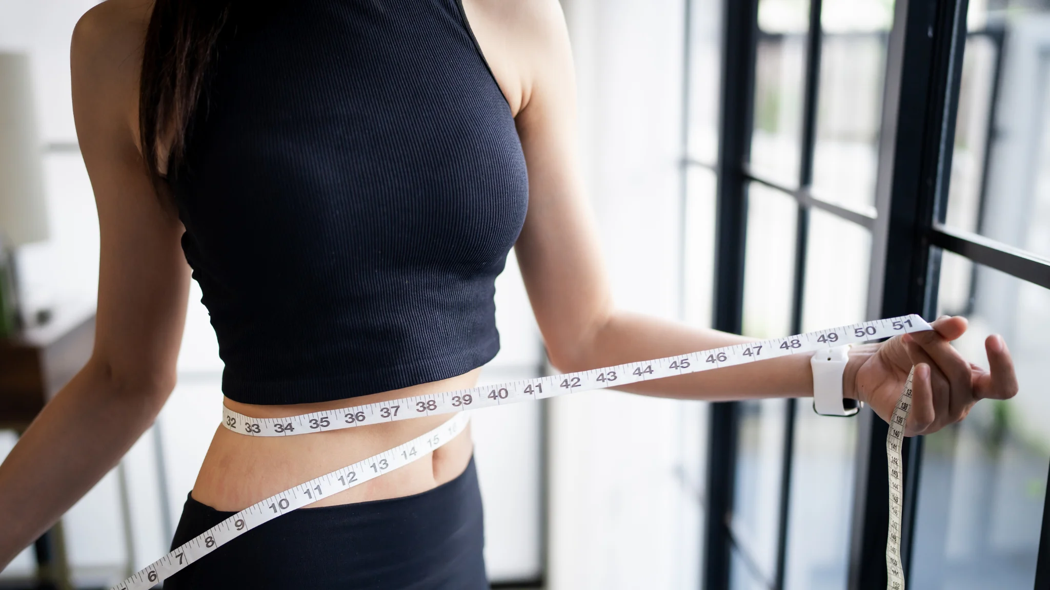 A woman in a black top measures her waist with a tape measure, focusing intently on her task.