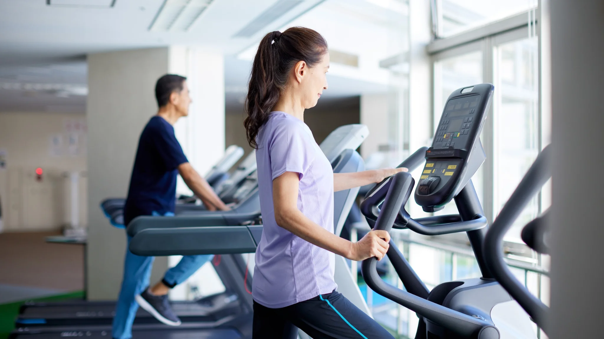 A woman and man running side by side on treadmills in a bright gym setting.