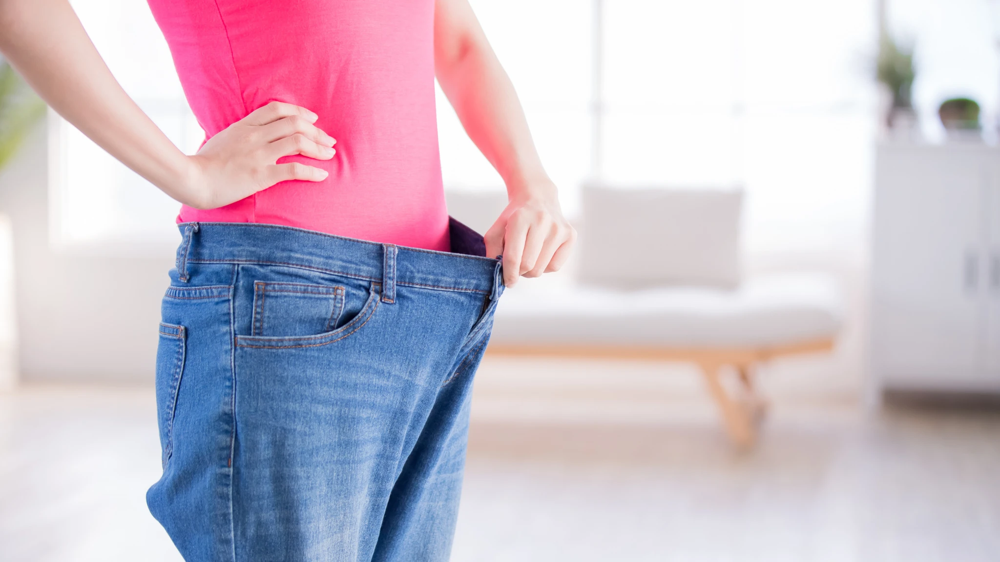 A woman stands with her hands on her waist, wearing fitted jeans and smiling confidently.