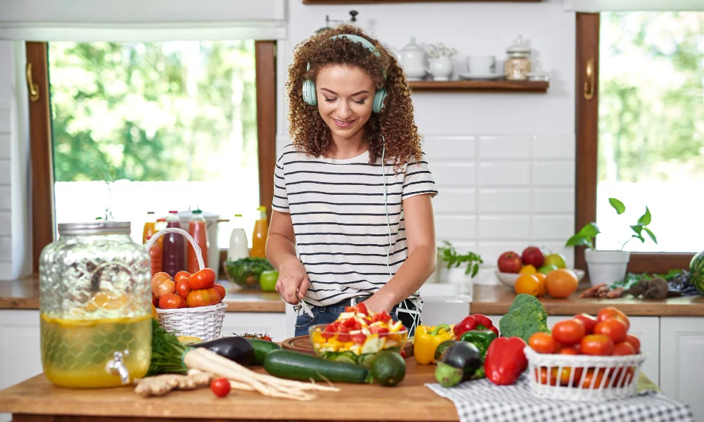 A woman with curly hair wearing headphones joyfully chops vegetables in a bright kitchen