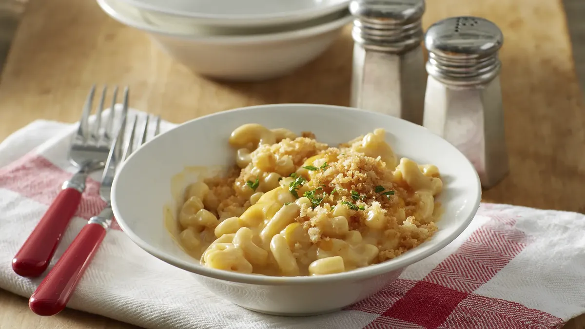 A bowl of creamy macaroni and cheese topped with toasted breadcrumbs and fresh chopped herbs, served on a red-and-white checkered napkin next to salt and pepper shakers.