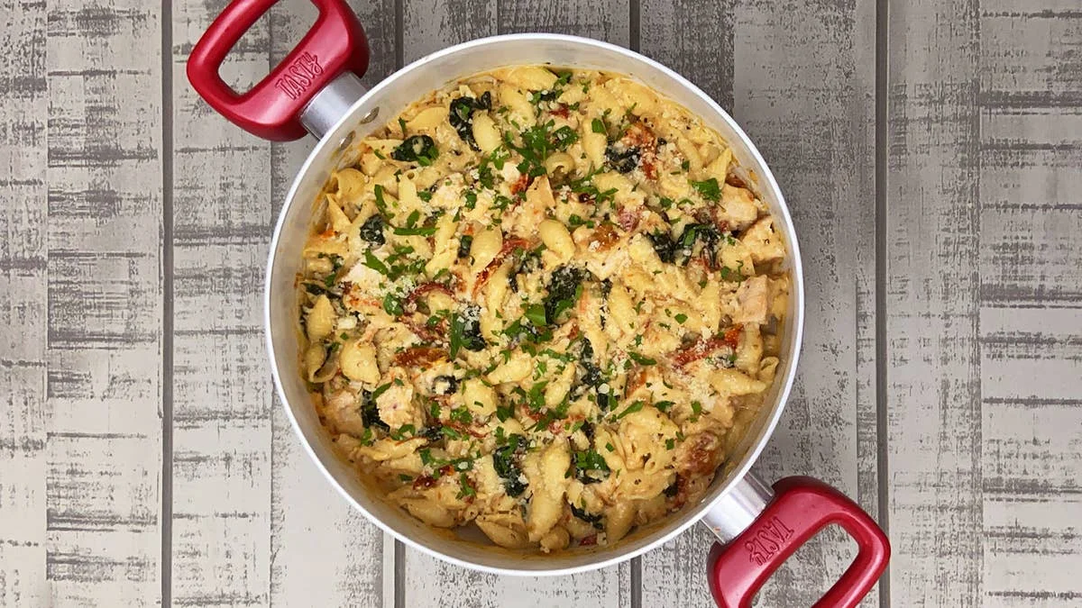 Top-down view of shell pasta tossed with chicken, spinach, and sun-dried tomatoes in a creamy sauce, garnished with fresh parsley and served in a pot with red handles.