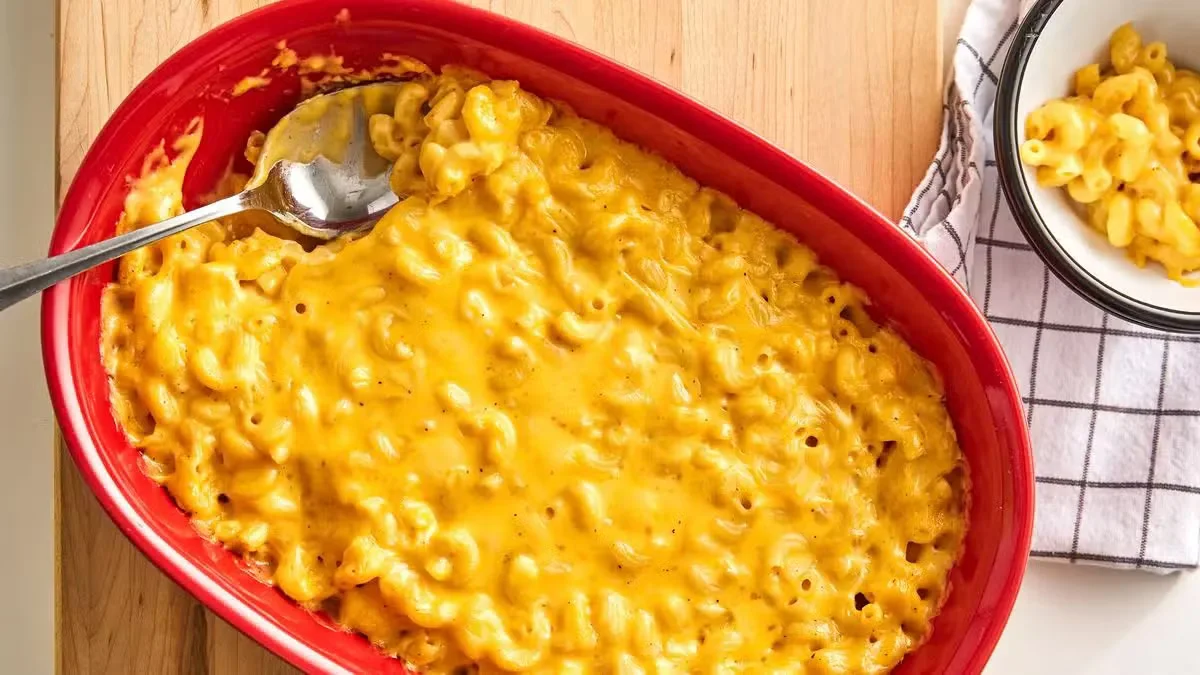 Baked macaroni and cheese with a smooth, melted cheddar cheese crust served in a red oval baking dish with a large metal serving spoon.