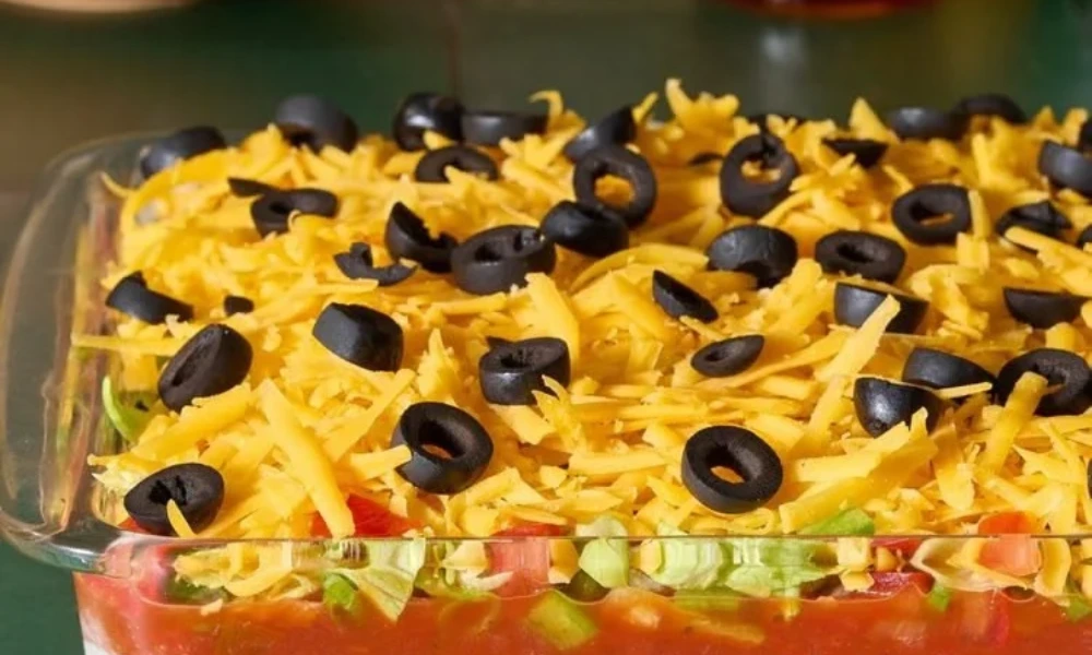 Layered 7-layer taco dip topped with shredded cheddar cheese, diced tomatoes, lettuce, and sliced black olives in a glass dish, perfect as a classic game day food appetizer.