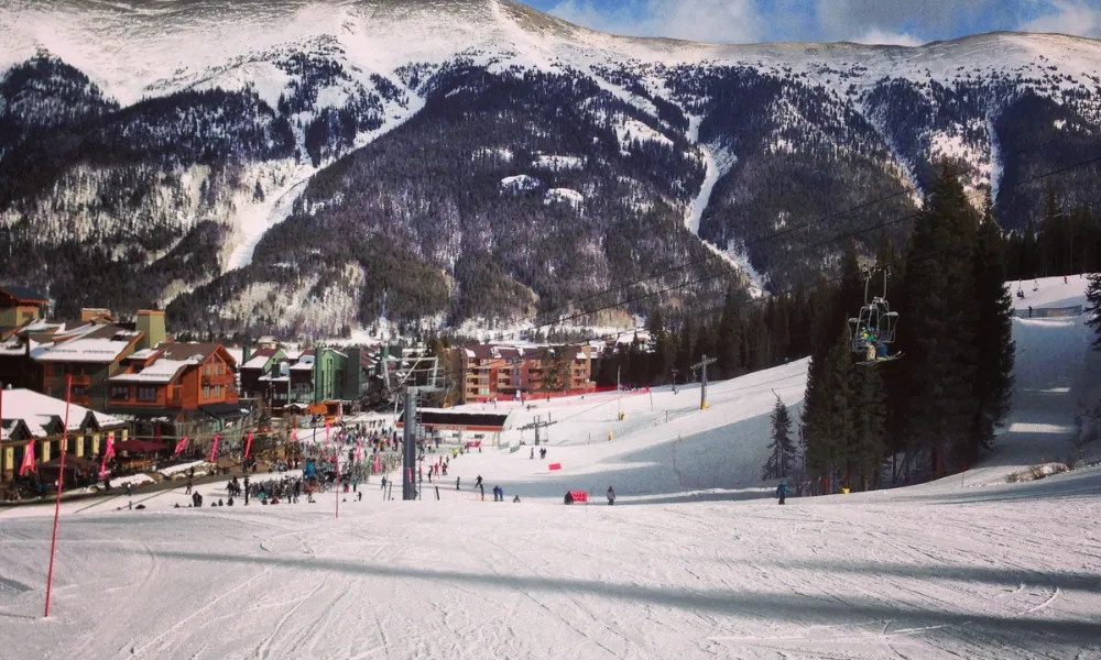 Best Ski Resorts in Colorado features a snow-covered ski slope leading down toward a mountain village with chairlifts operating against a backdrop of alpine peaks.