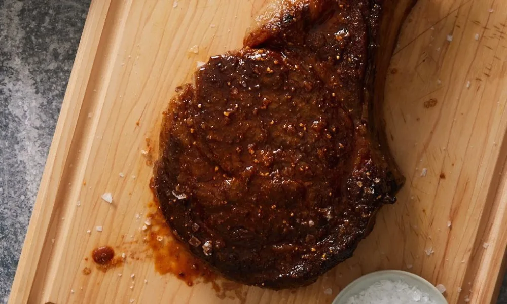Thick juicy grilled cowboy steak seasoned with flaky salt on a wooden board. 