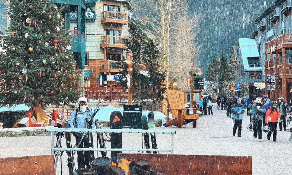 Best Ski Resorts in Colorado features a snow-dusted ski village plaza complete with an outdoor log fire pit, decorated Christmas tree, and visitors walking past alpine lodges.