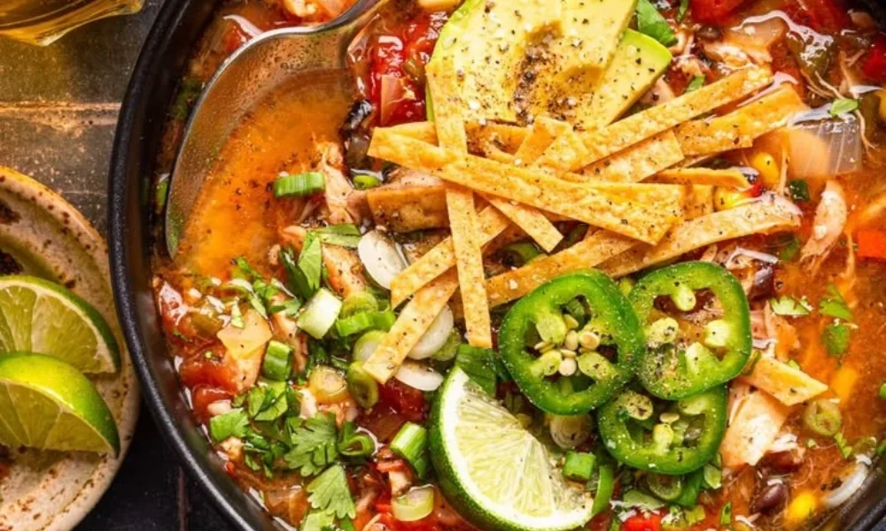 A bowl of chicken tortilla soup topped with crispy tortilla strips, sliced avocado, jalapeños, cilantro, green onions, and fresh lime, perfect for comforting mexican dinner recipes. 