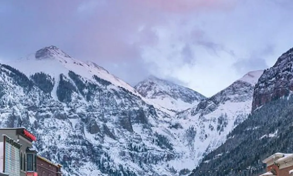 Best Ski Resorts in Colorado showcased by a picturesque view down a snow-covered historic mountain town main street framed by towering snow-capped peaks at dusk.