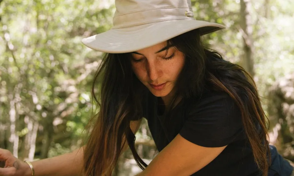 Best Hiking Hats demonstrated by a woman wearing a cream wide-brim sun hat with top mesh ventilation outdoors among trees.