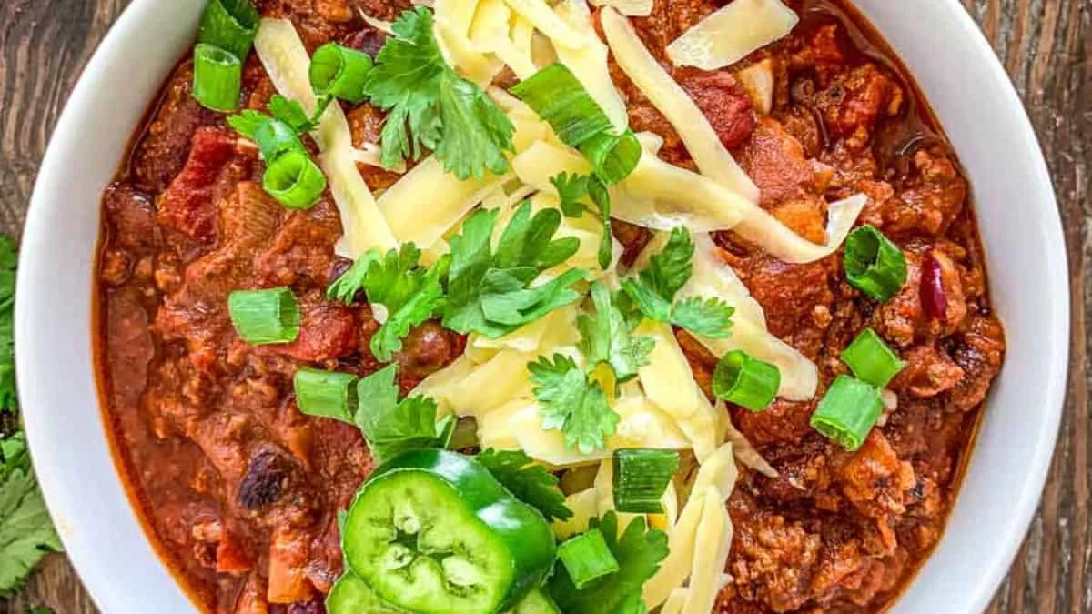 A white bowl of hearty beef chili topped with shredded cheese, chopped green onions, fresh cilantro, and jalapeño slices.