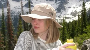 Best Hiking Hats worn by a hiker with a beige brimmed bucket hat, holding a yellow water bottle against a backdrop of snow-dusted mountains.