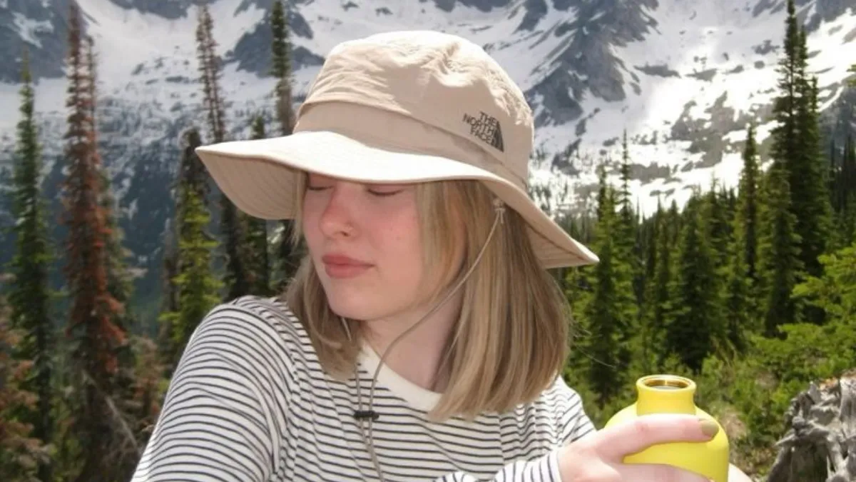Best Hiking Hats worn by a hiker with a beige brimmed bucket hat, holding a yellow water bottle against a backdrop of snow-dusted mountains.