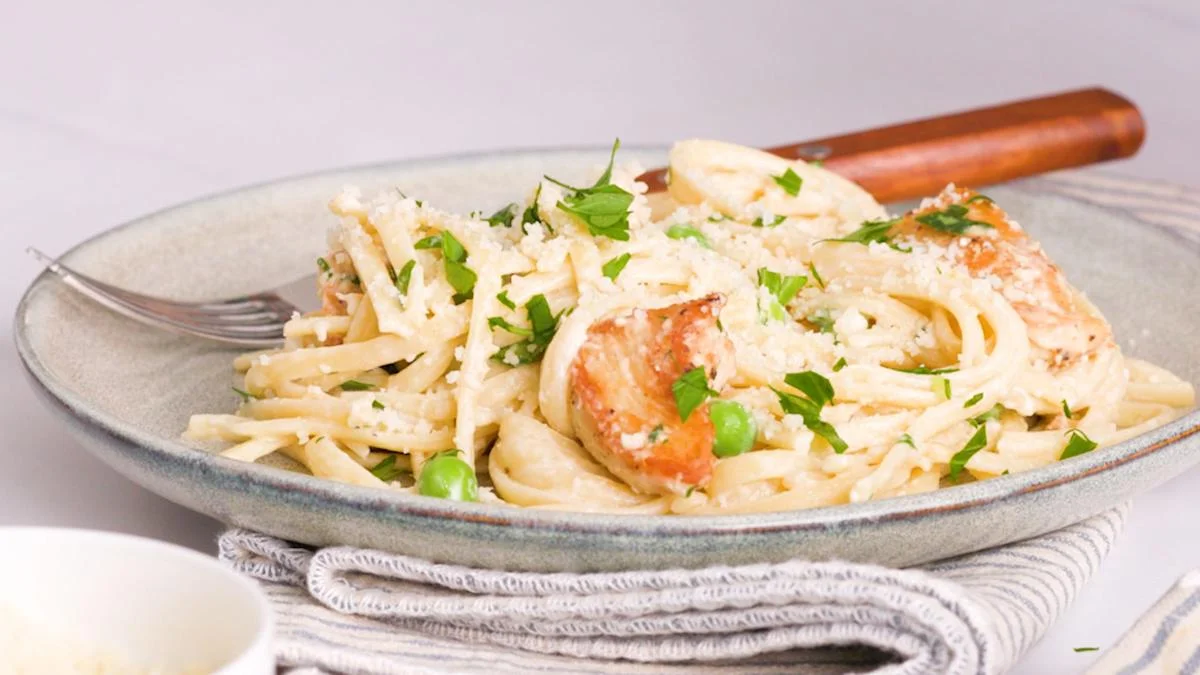 Creamy chicken fettuccine pasta topped with green peas, grated Parmesan cheese, and fresh chopped parsley, served on a grey plate with a fork.