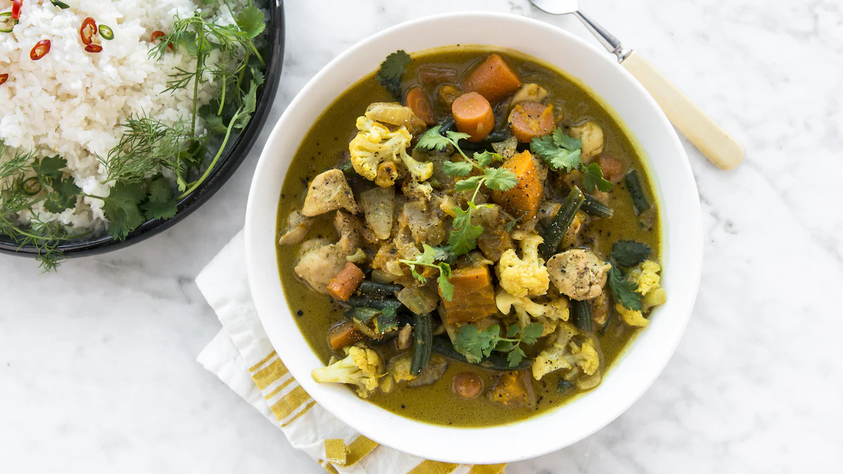 A bowl of Thai green curry packed with cauliflower florets, carrots, sweet potatoes, and green beans, served alongside a side of white steamed rice garnished with cilantro and sliced chili peppers.